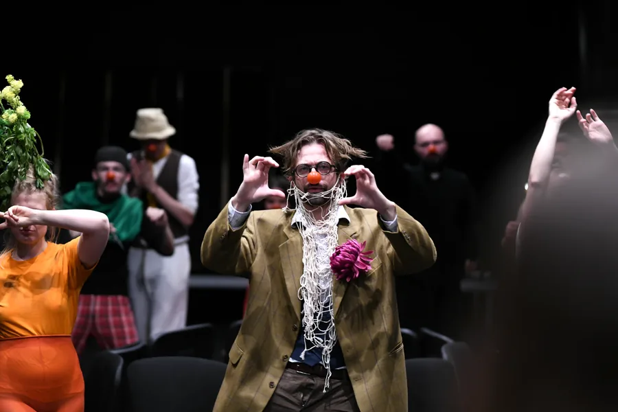 A performer with a beard and a flower gesturing towards the audience.