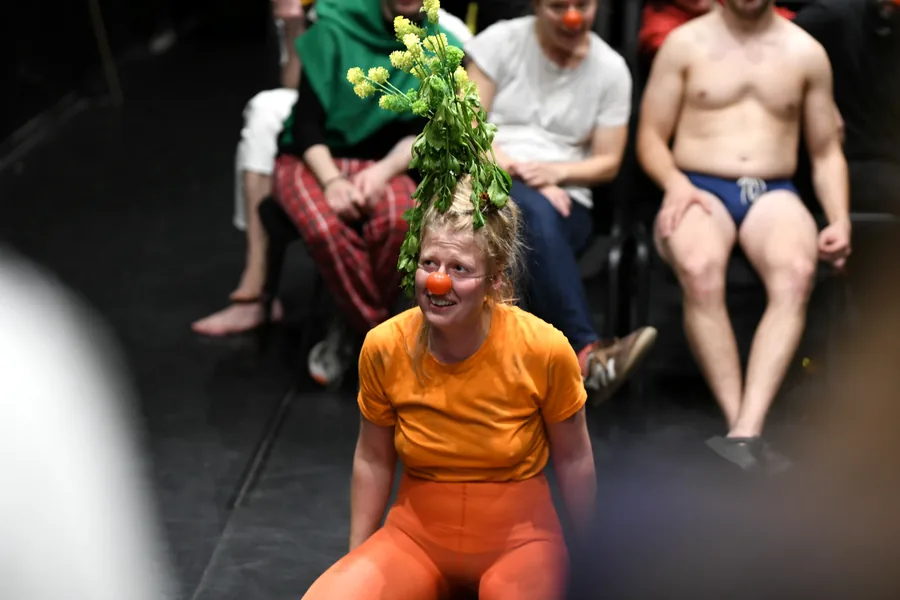 A performer in orange with a leafy headpiece, surrounded by the cast.